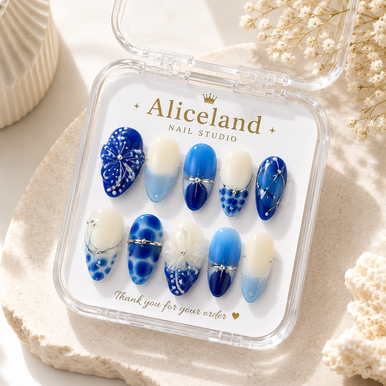 A set of 10 press-on nails in a clear plastic case with the brand name 'Aliceland Nail Studio' printed on the insert. The nails feature various blue and white designs including gradients, splatters, and glittery details, styled on a beige stone surface with dried flowers.