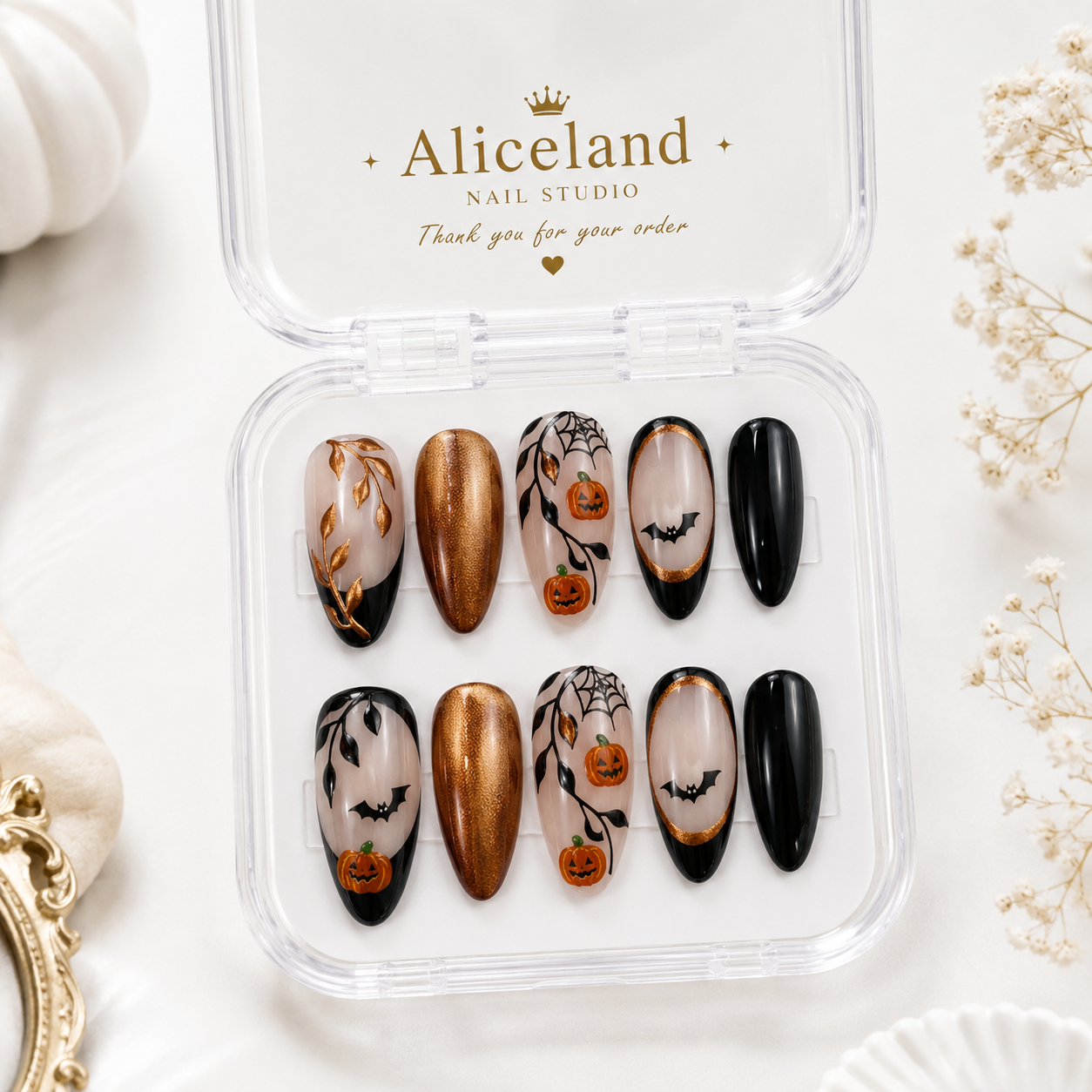 A set of Halloween-themed press-on nails in a clear plastic case with the brand name 'Aliceland Nail Studio' printed on the inside lid. The nails feature designs of pumpkins, bats, spiderwebs, and gold metallic accents on a white background.