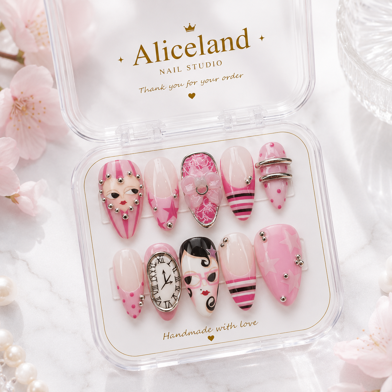 A set of 10 pink and white decorative press-on nails displayed in a clear plastic case with the brand name 'Aliceland NAIL STUDIO' printed on the inside lid. The nails feature various designs including polka dots, stripes, rhinestones, and face illustrations.