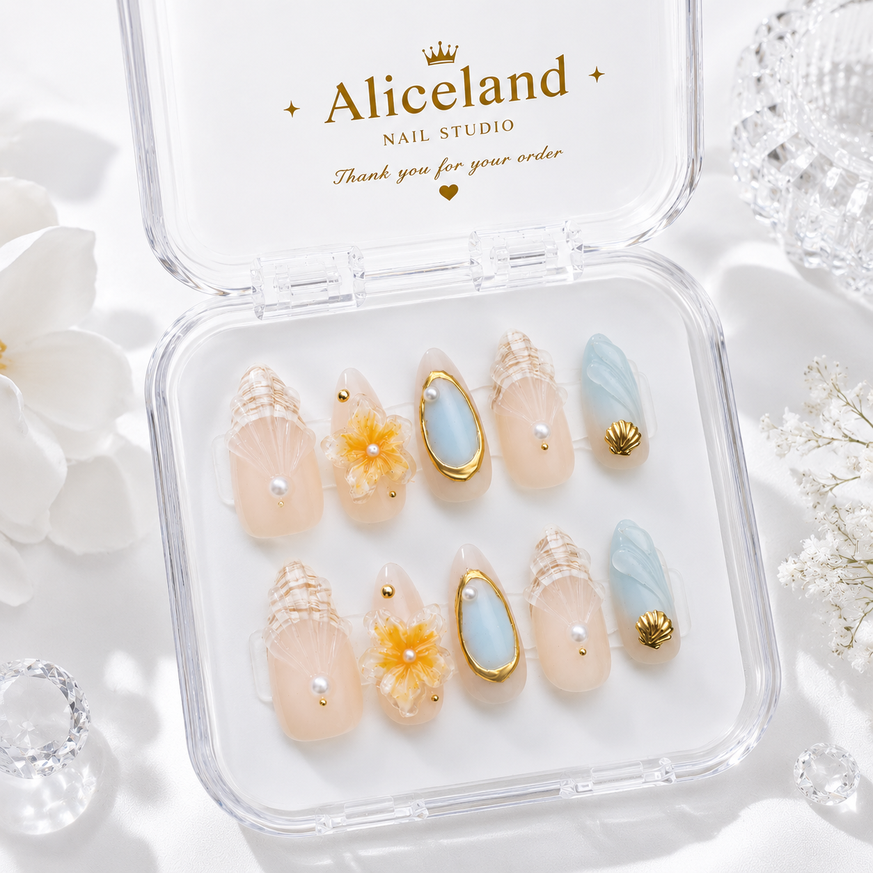 A clear plastic case of Aliceland nail studio press-on false nails displayed open on a white background with decorative white flowers and crystal props. The nails feature beige and peach tones with shell, pearl, yellow flower, light blue, and gold shell designs.